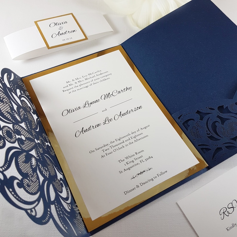 May include: A navy blue and gold wedding invitation with a laser-cut design. The invitation reads "Olivia Lynne McCarthy and Andrew Lee Anderson" and includes the date and time of the wedding. The invitation also includes the location of the wedding, "The White Room, 1 King Street, St Augustine, FL 32084".