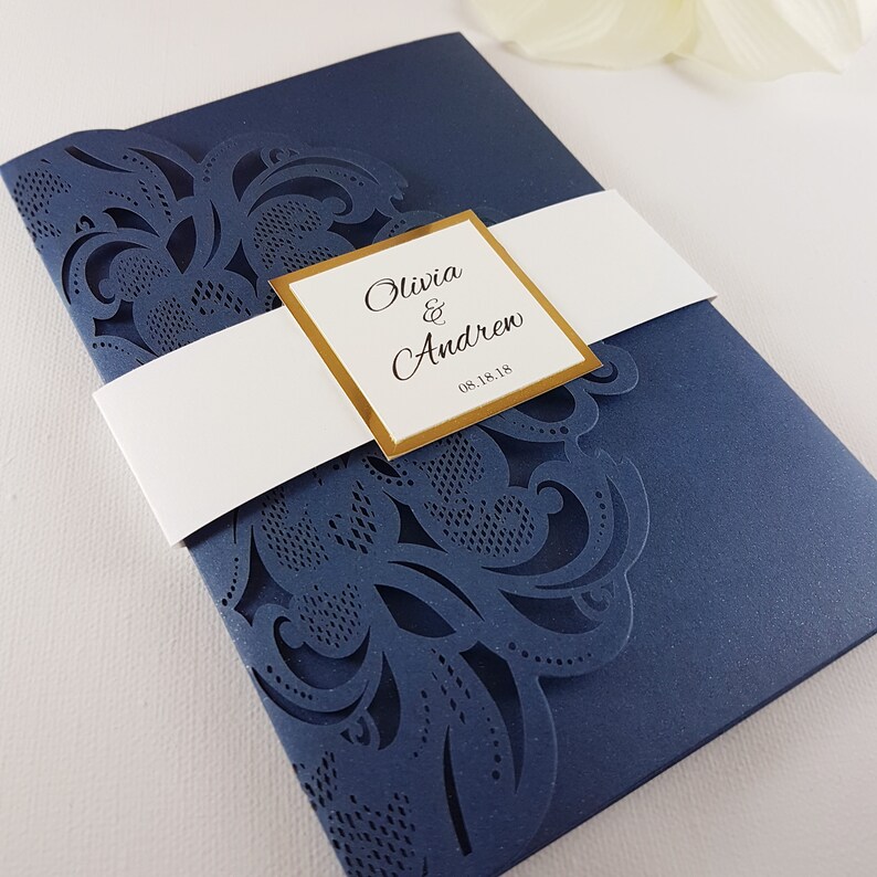 May include: Navy blue wedding invitation with a white satin ribbon and a gold square label with the text "Olivia & Andrew 08.18.18". The invitation features a laser-cut design with intricate floral patterns.