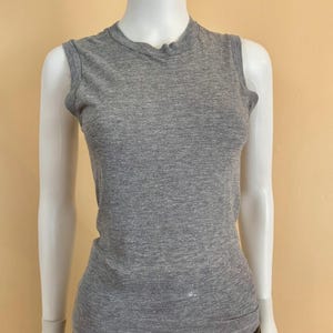 May include: A sleeveless grey heathered tank top with a crew neckline, featuring a fitted silhouette and a straight hemline. The garment is displayed on a mannequin, showcasing the textured knit fabric and the simple, clean design of the top.