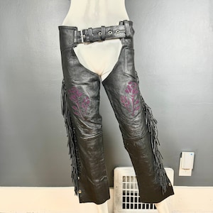 May include: Black leather chaps with fringe and purple floral embroidery. The chaps have a belt with a silver buckle.