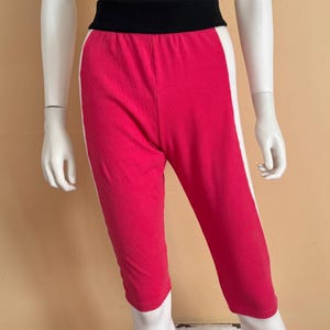 May include: Capri-length leggings featuring a vibrant hot pink main body, a wide black elastic waistband, and contrasting white vertical side stripes running down the outer legs. The garment is displayed on a mannequin torso and upper legs.