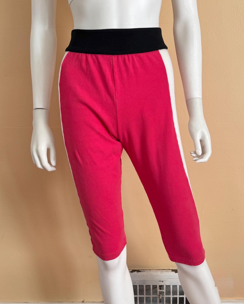 May include: Capri-length leggings featuring a vibrant hot pink main body, a wide black elastic waistband, and contrasting white vertical side stripes running down the outer legs. The garment is displayed on a mannequin torso and upper legs.