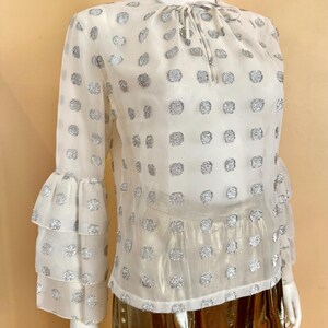 Op de afbeelding: Een doorschijnende witte blouse met zilveren stippen en lange mouwen met gelaagde ruches. De blouse heeft een strik bij de halslijn. Het kledingstuk wordt op een mannequin getoond.