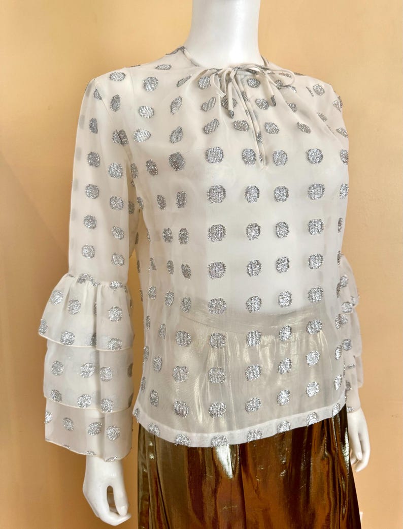 Op de afbeelding: Een doorschijnende witte blouse met zilveren stippen en lange mouwen met gelaagde ruches. De blouse heeft een strik bij de halslijn. Het kledingstuk wordt op een mannequin getoond.