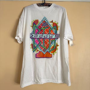 May include: White t-shirt with a colorful graphic design. The design features the text "DADDY'S BIG SURF SHOP" in a geometric, retro style, with vibrant colors including orange, pink, green, and blue. The shirt is hanging on a black hanger.