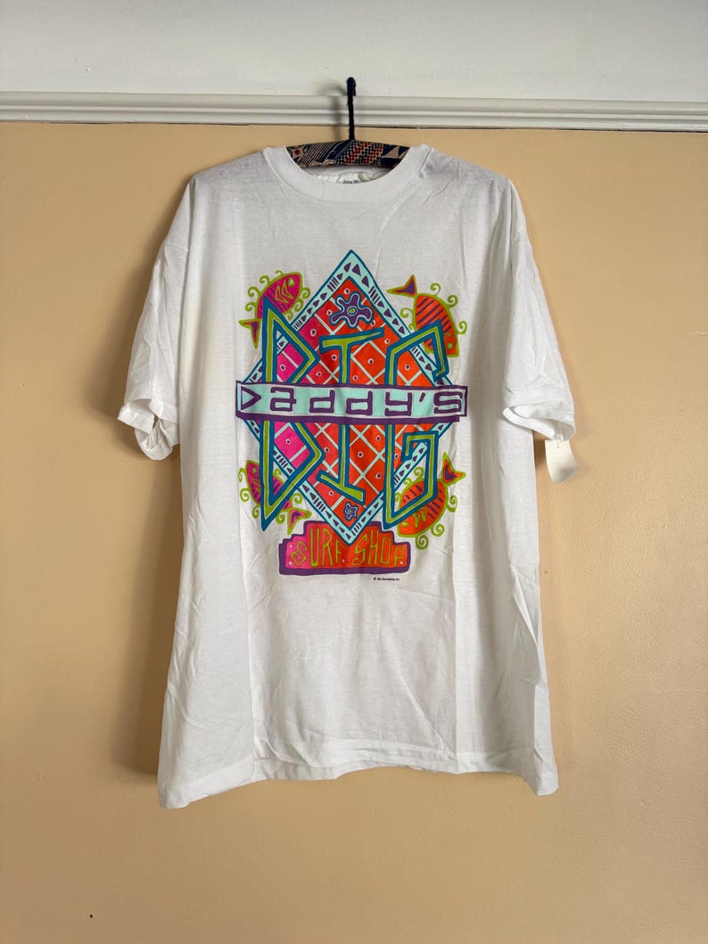 May include: White t-shirt with a colorful graphic design. The design features the text "DADDY'S BIG SURF SHOP" in a geometric, retro style, with vibrant colors including orange, pink, green, and blue. The shirt is hanging on a black hanger.