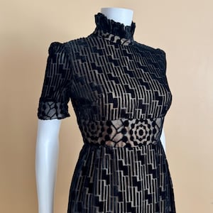 May include: Black vintage dress with a high ruffled collar and short sleeves. The dress features a geometric pattern and a decorative waist panel. The dress is displayed on a white mannequin.