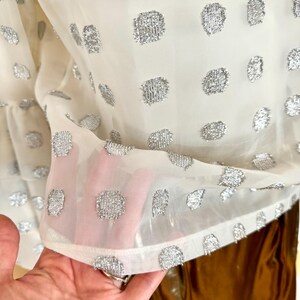 Op de afbeelding: Een doorschijnende, cr&egrave;mekleurige blouse met een patroon van zilveren, getextureerde stippen. De stof is licht en enigszins transparant, met een gefranjerde zoom. De blouse is een stijlvol kledingstuk.
