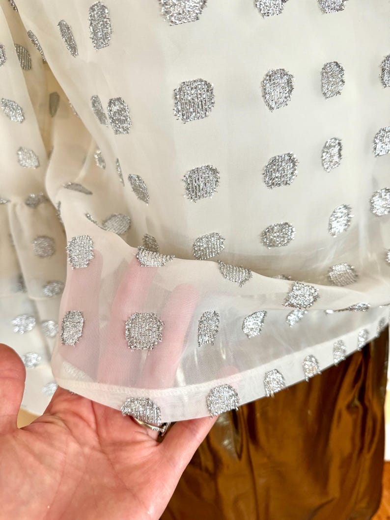 Op de afbeelding: Een doorschijnende, cr&egrave;mekleurige blouse met een patroon van zilveren, getextureerde stippen. De stof is licht en enigszins transparant, met een gefranjerde zoom. De blouse is een stijlvol kledingstuk.