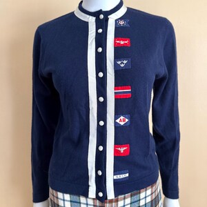 May include: A navy blue cardigan with white trim and a row of buttons down the front. The cardigan has several embroidered patches with red, white, and blue designs, including a flag, an eagle, and the letters "AB". The cardigan also has a small patch with the text "NASM".