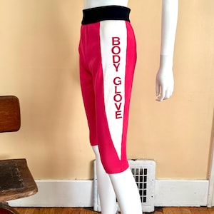 May include: Hot pink knee-length shorts featuring a wide black waistband and a vertical white side panel. The white panel displays the text BODY GLOVE in red capital letters. The shorts are displayed on a white mannequin torso and legs.