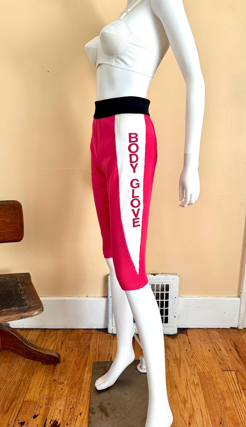 May include: Hot pink knee-length shorts featuring a wide black waistband and a vertical white side panel. The white panel displays the text BODY GLOVE in red capital letters. The shorts are displayed on a white mannequin torso and legs.