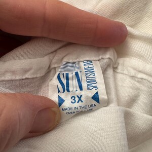 May include: A white t-shirt tag with the text "SUN SPORTSWEAR" and "3X" in blue. The tag also reads "MADE IN THE USA" and "OVER FOR CARE". The tag is held by a person's fingers.