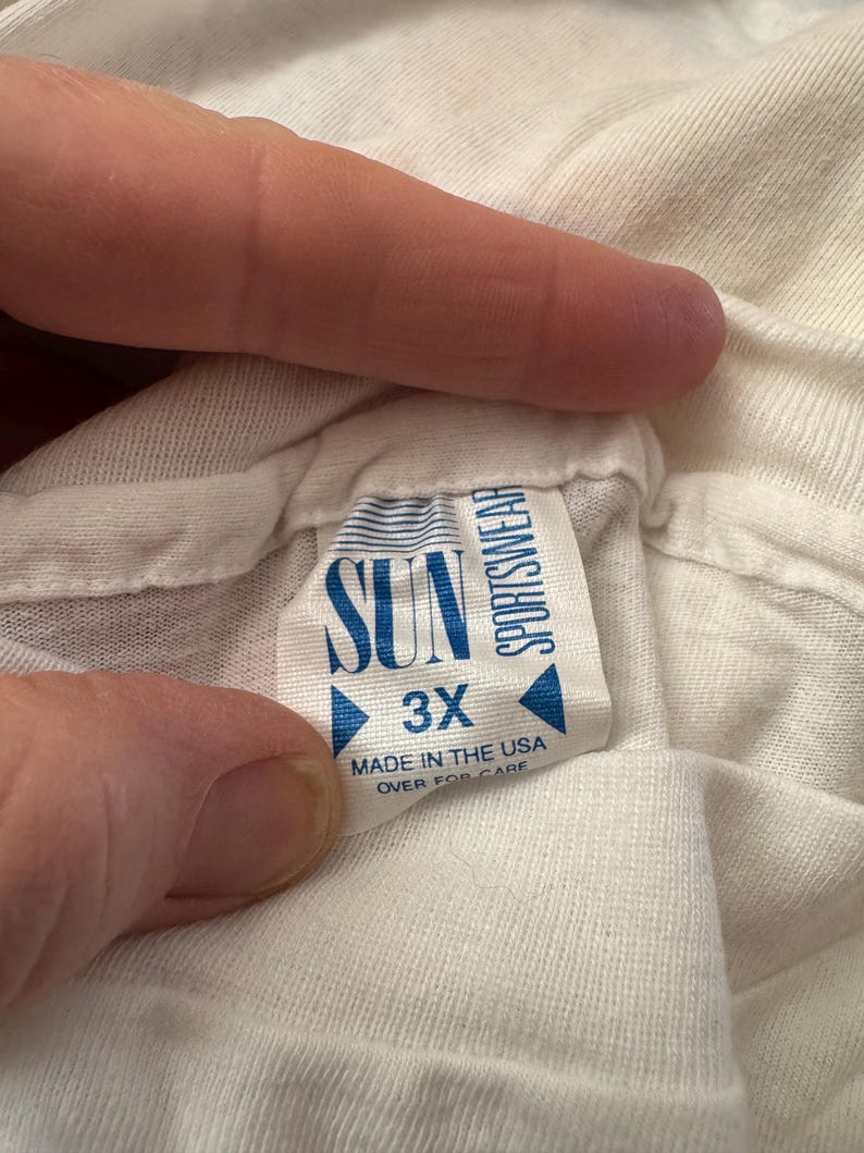 May include: A white t-shirt tag with the text "SUN SPORTSWEAR" and "3X" in blue. The tag also reads "MADE IN THE USA" and "OVER FOR CARE". The tag is held by a person's fingers.