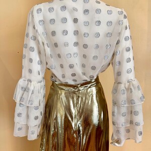 Op de afbeelding: Een witte blouse met zilveren stippen en lange mouwen met ruches. De blouse wordt gecombineerd met een gouden metallic rok. De outfit wordt gepresenteerd op een mannequin.