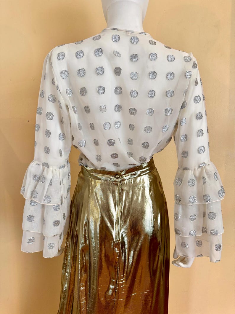 Op de afbeelding: Een witte blouse met zilveren stippen en lange mouwen met ruches. De blouse wordt gecombineerd met een gouden metallic rok. De outfit wordt gepresenteerd op een mannequin.