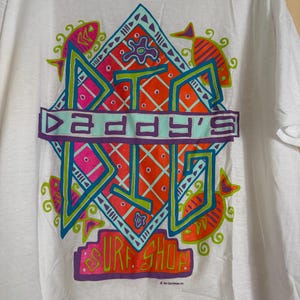 May include: White t-shirt featuring a colorful graphic design. The design includes the text "DADDY'S" in a turquoise banner, surrounded by geometric shapes, fish, and the words "SURF SHOP" in bright colors. A vintage surf shop tee.