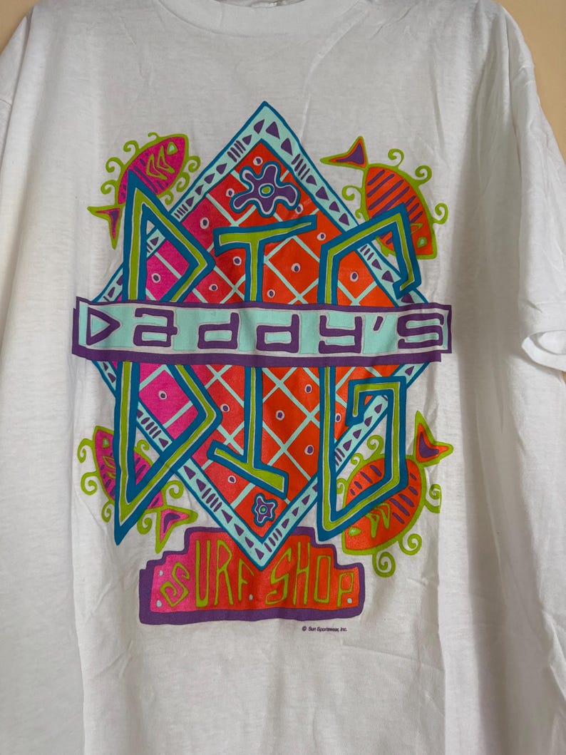 May include: White t-shirt featuring a colorful graphic design. The design includes the text "DADDY'S" in a turquoise banner, surrounded by geometric shapes, fish, and the words "SURF SHOP" in bright colors. A vintage surf shop tee.