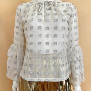 Op de afbeelding: Een doorschijnende witte blouse met zilveren stippen en gelaagde, gerimpelde mouwen. De blouse heeft een strik bij de halslijn en wordt gecombineerd met een gouden metallic rok. De top wordt op een mannequin getoond.