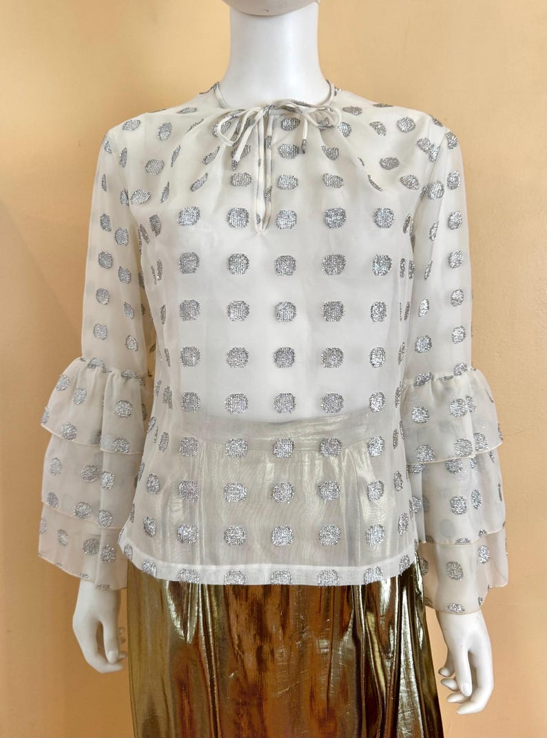 Op de afbeelding: Een doorschijnende witte blouse met zilveren stippen en gelaagde, gerimpelde mouwen. De blouse heeft een strik bij de halslijn en wordt gecombineerd met een gouden metallic rok. De top wordt op een mannequin getoond.