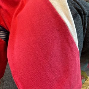 May include: A knit garment featuring a vibrant red section adjacent to a solid white stripe. The fabric displays a fine, consistent texture throughout the red and white panels, with a visible seam line running along the edge of the red fabric.