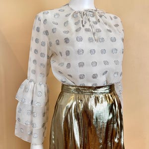 Op de afbeelding: Een witte blouse met zilveren stippen en gelaagde, gerimpelde mouwen, gecombineerd met een metallic gouden rok. De blouse heeft een strikdetail in de hals. De rok heeft een hoge taille en een glanzend, reflecterend oppervlak.