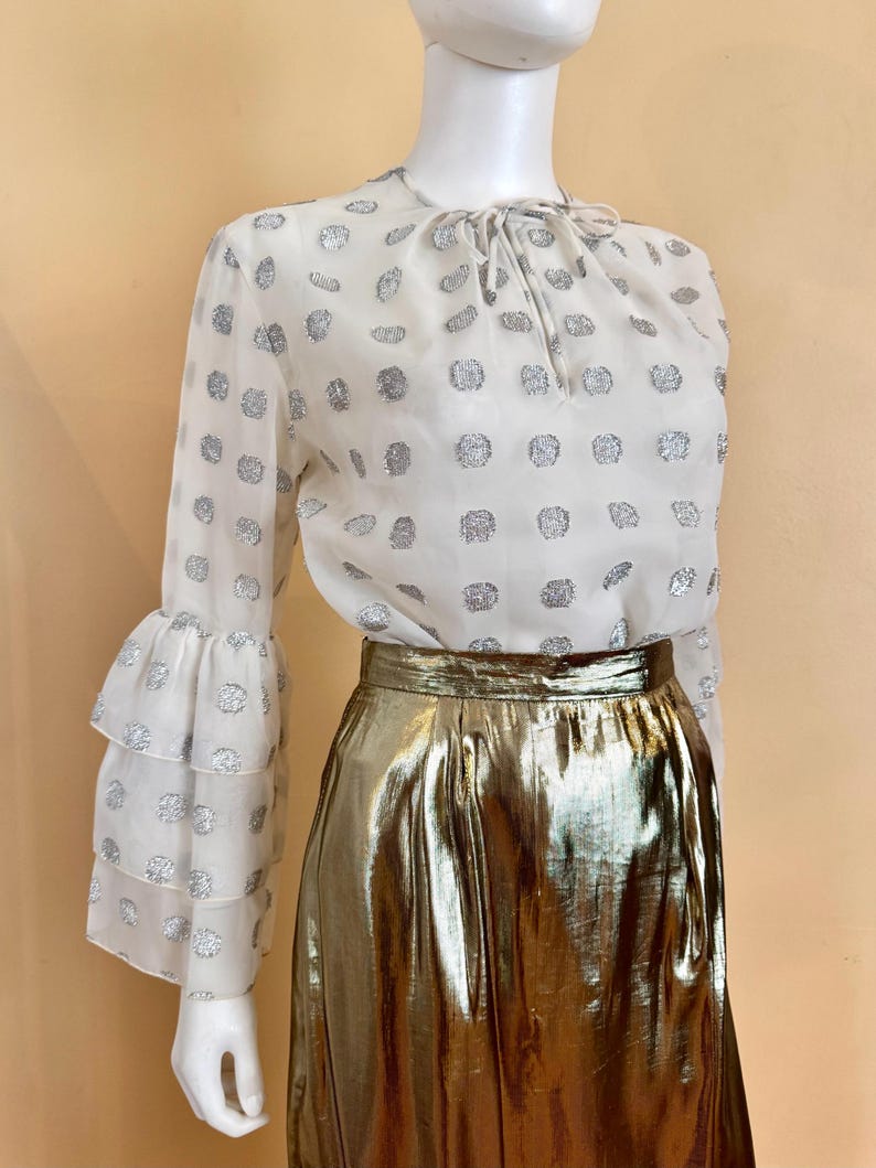 Op de afbeelding: Een witte blouse met zilveren stippen en gelaagde, gerimpelde mouwen, gecombineerd met een metallic gouden rok. De blouse heeft een strikdetail in de hals. De rok heeft een hoge taille en een glanzend, reflecterend oppervlak.