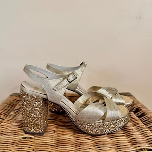 May include: A pair of gold glitter platform sandals with a criss-cross strap design. The sandals have a chunky heel and a buckle closure on the ankle strap. The platform measures approximately 1.5 inches, and the heel is about 4 inches.