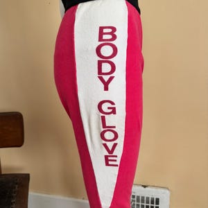 May include: Hot pink leggings featuring a wide white vertical side panel with the text BODY GLOVE printed in dark red letters. The leggings have a thick black waistband and a fitted silhouette that extends to the mid-calf area of the leg.