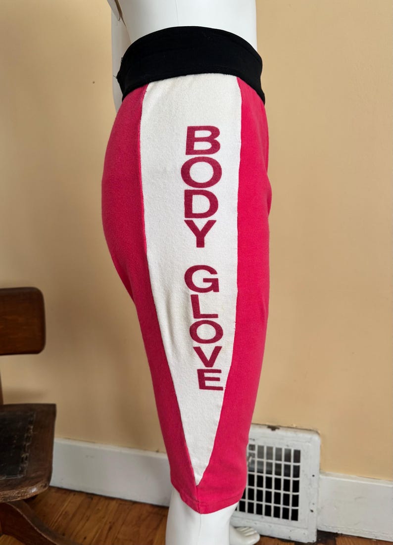 May include: Hot pink leggings featuring a wide white vertical side panel with the text BODY GLOVE printed in dark red letters. The leggings have a thick black waistband and a fitted silhouette that extends to the mid-calf area of the leg.