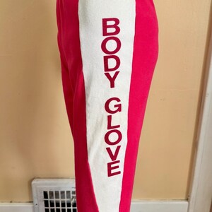 May include: Bright pink athletic pants featuring a wide black waistband and a vertical white side panel. The text BODY GLOVE is printed in a bold, dark red font vertically along the white panel on the side of the leg.