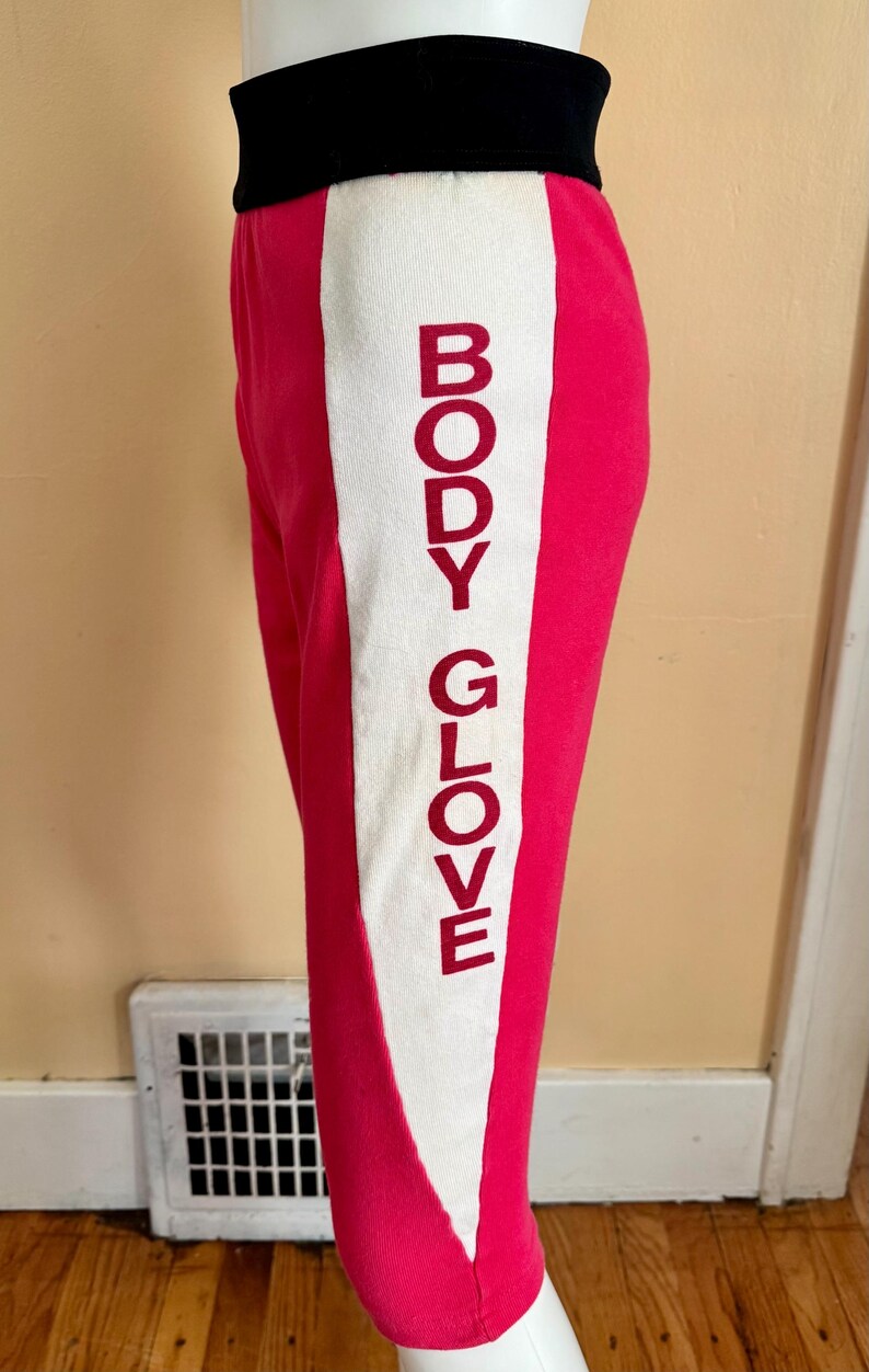 May include: Bright pink athletic pants featuring a wide black waistband and a vertical white side panel. The text BODY GLOVE is printed in a bold, dark red font vertically along the white panel on the side of the leg.