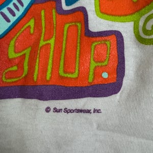 May include: Close-up of a white t-shirt with a colorful graphic design. The design features the word "SHOP" in orange with a green outline, surrounded by blue, green, and purple shapes. The text "Sun Sportswear, Inc." is printed below the graphic.