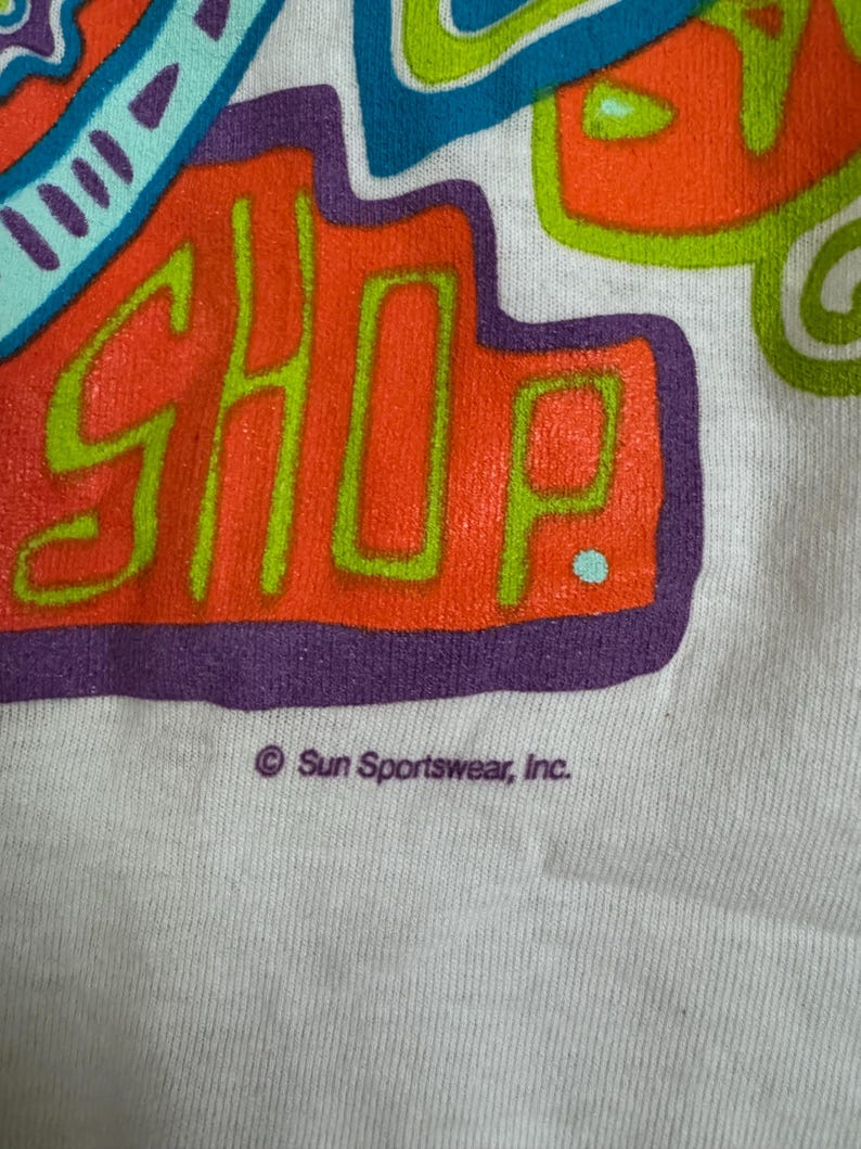 May include: Close-up of a white t-shirt with a colorful graphic design. The design features the word "SHOP" in orange with a green outline, surrounded by blue, green, and purple shapes. The text "Sun Sportswear, Inc." is printed below the graphic.