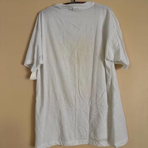 May include: A white, short-sleeved t-shirt hanging on a black hanger. The shirt is plain and appears to be made of a lightweight fabric. A price tag is attached to the shirt. The shirt is hanging against a light tan wall.