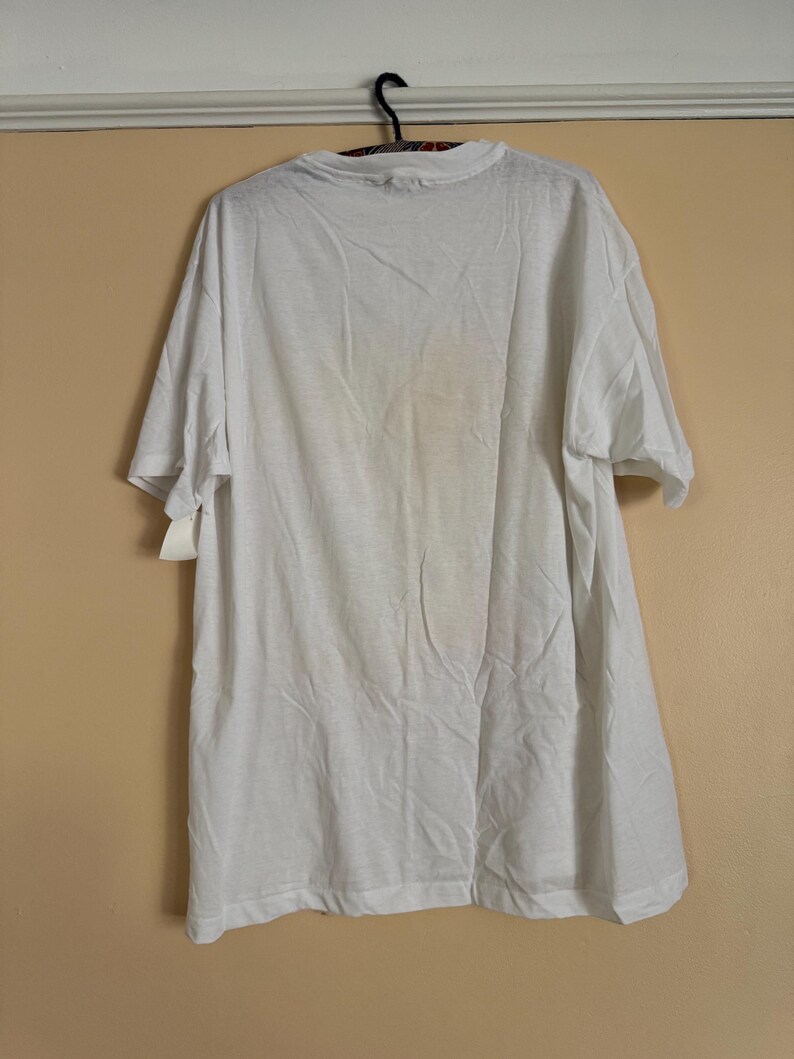May include: A white, short-sleeved t-shirt hanging on a black hanger. The shirt is plain and appears to be made of a lightweight fabric. A price tag is attached to the shirt. The shirt is hanging against a light tan wall.