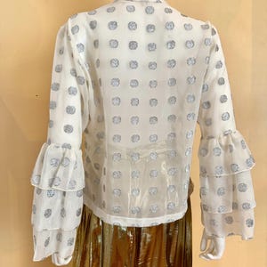 Op de afbeelding: Witte blouse met zilveren stippen en gelaagde ruchesmouwen. De blouse wordt gecombineerd met een gouden metallic rok. Het kledingstuk wordt gepresenteerd op een mannequin tegen een lichtgele achtergrond.
