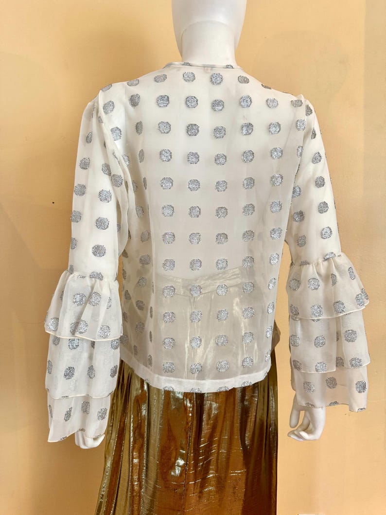 Op de afbeelding: Witte blouse met zilveren stippen en gelaagde ruchesmouwen. De blouse wordt gecombineerd met een gouden metallic rok. Het kledingstuk wordt gepresenteerd op een mannequin tegen een lichtgele achtergrond.