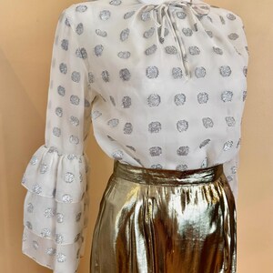 Op de afbeelding: Een witte blouse met zilveren stippen en lange mouwen met ruches manchetten wordt gecombineerd met een gouden metallic rok. De blouse heeft een strik bij de halslijn. De rok heeft een midi-lengte.