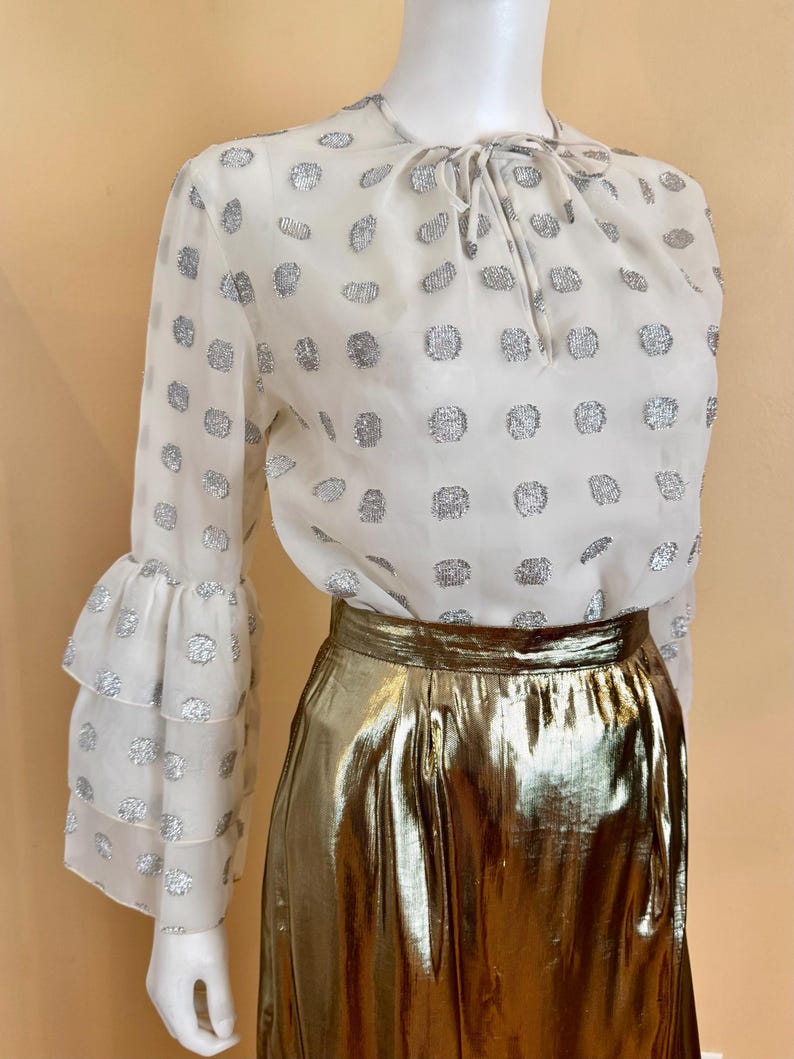 Op de afbeelding: Een witte blouse met zilveren stippen en lange mouwen met ruches manchetten wordt gecombineerd met een gouden metallic rok. De blouse heeft een strik bij de halslijn. De rok heeft een midi-lengte.