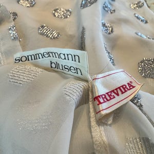 Op de afbeelding: Close-up van een witte blouse met zilveren glitteraccenten. De blouse heeft een delicate, transparante stof en is versierd met verschillende vormen van zilveren glitter. De labels lezen "sommermann blusen" en "TREVIRA".