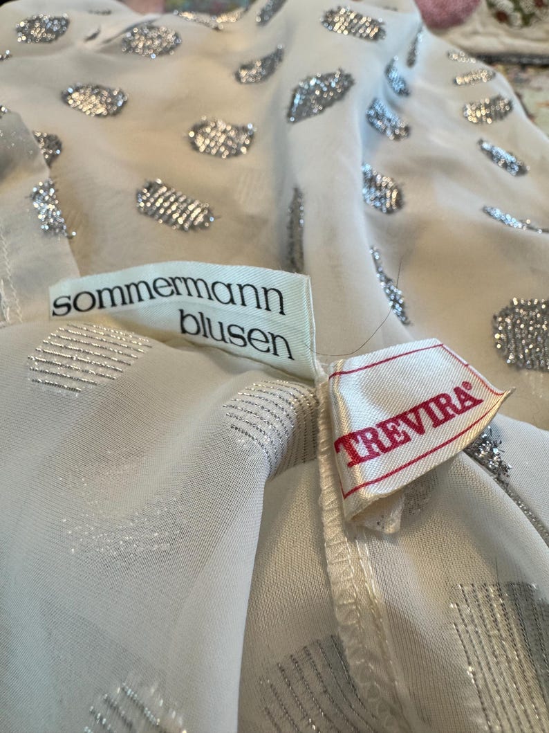 Op de afbeelding: Close-up van een witte blouse met zilveren glitteraccenten. De blouse heeft een delicate, transparante stof en is versierd met verschillende vormen van zilveren glitter. De labels lezen "sommermann blusen" en "TREVIRA".