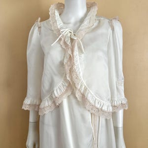 May include: A white satin robe set featuring a bolero-style jacket with three-quarter length sleeves, ruffled edges, and beige lace trim. The jacket includes a front tie closure. The matching sleeveless robe has a wrap-style front with lace trim.