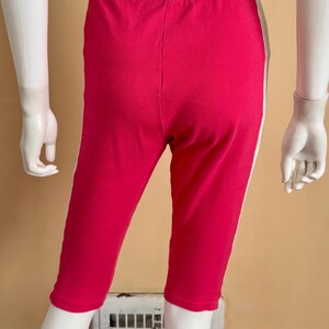 May include: Bright pink capri leggings featuring a wide, solid black elastic waistband and a vertical white stripe running down each outer side. The garment reaches mid-calf length and is displayed on a mannequin to show the fit and design.