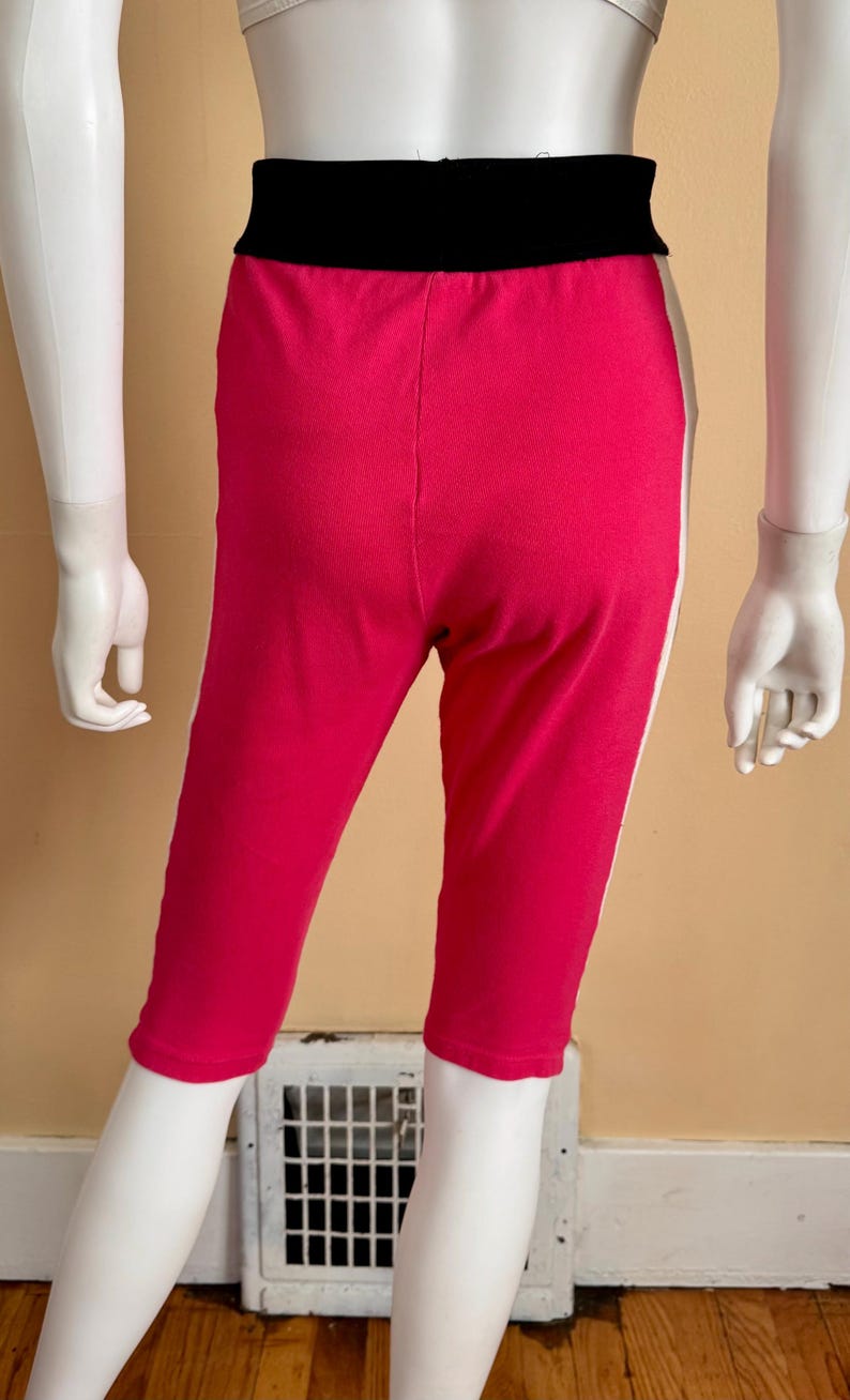 May include: Bright pink capri leggings featuring a wide, solid black elastic waistband and a vertical white stripe running down each outer side. The garment reaches mid-calf length and is displayed on a mannequin to show the fit and design.