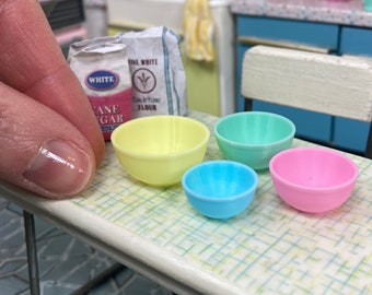 Puede incluir: Cuatro boles de mezcla en miniatura en amarillo, azul claro, verde y rosa. Los boles están sobre una superficie a cuadros blancos y verdes. Una bolsa de harina blanca está en el fondo.