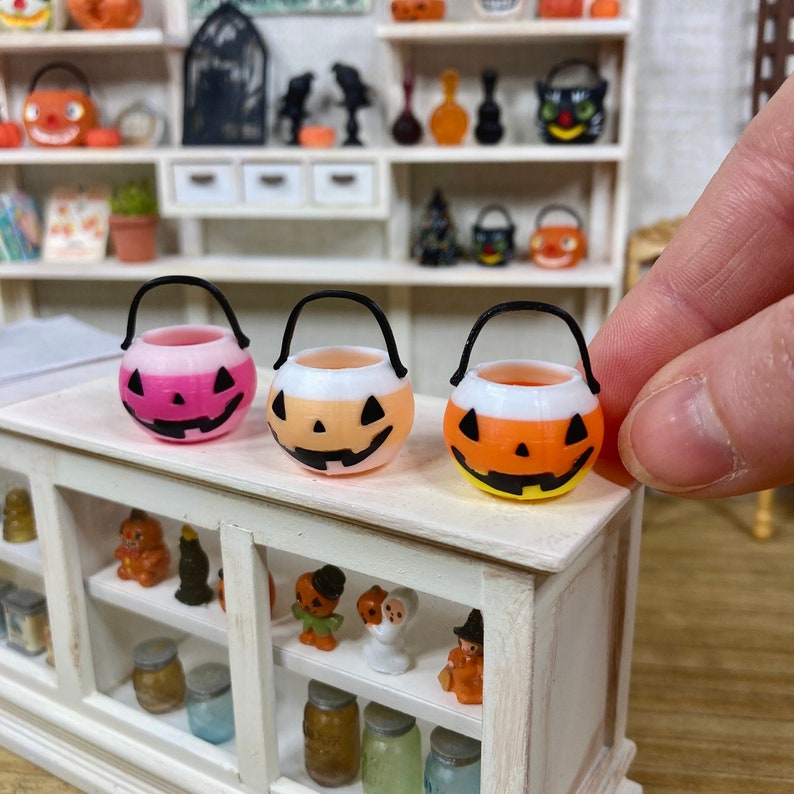 May include: Three miniature plastic Halloween trick-or-treat buckets. The buckets are shaped like jack-o'-lanterns with black eyes, noses, and mouths. The buckets are pink, orange, and orange and white.