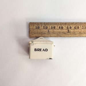 May include: A miniature cream-colored metal bread box with a lid and the word BREAD printed in black on the front. A wooden ruler is positioned above the item, showing it measures approximately 1.5 inches in width at its widest point.