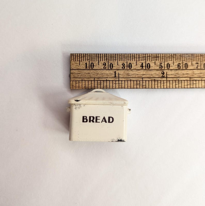May include: A miniature cream-colored metal bread box with a lid and the word BREAD printed in black on the front. A wooden ruler is positioned above the item, showing it measures approximately 1.5 inches in width at its widest point.