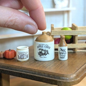 May include: Miniature ceramic apple cider and pumpkin themed decor. The set includes a small jug with a handle, a bottle, and a jar, all with labels. A tiny orange pumpkin is also present. The jug reads "Apple Cider" and "Farm Fresh".